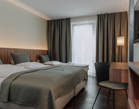 Modern hotel room with two beds, a desk and a chair, a large window with sheer and dark curtains and soft lighting.