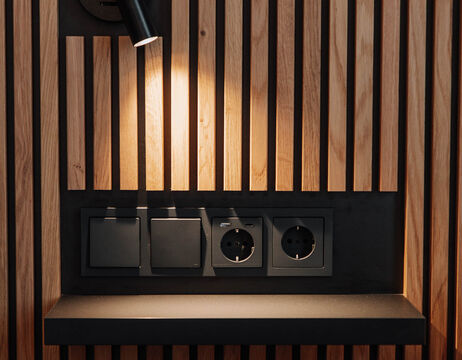 A spotlight illuminates sockets and switches on a wooden slatted wall above a dark shelf.