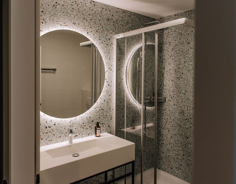 Modernes Badezimmer mit Terrazzo-Wänden, einem runden, hinterleuchteten Spiegel, Waschbecken und einer Glasduschkabine.