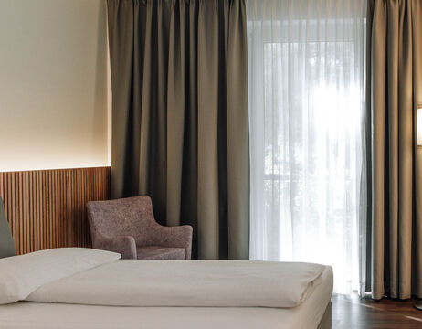 Ein Hotelzimmer mit einem ordentlich gemachten Bett, einem malvenfarbenen Sessel und großen Fenstern mit durchsichtigen Vorhängen.