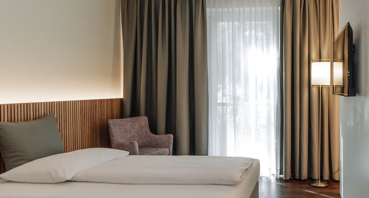 Minimalist hotel room with a bed, a gray chair, a floor lamp, a wall-mounted TV and a large window with curtains.