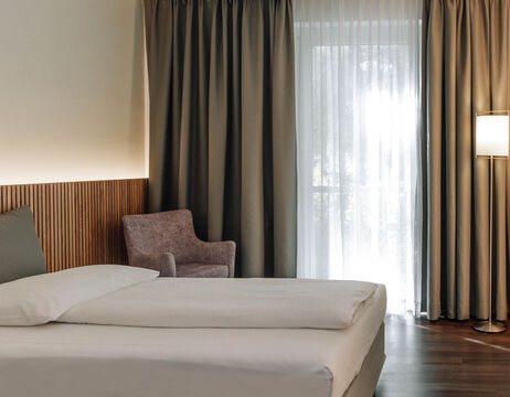 Minimalistisches Hotelzimmer mit einem ordentlich gemachten Bett, einem Sessel, einer Stehlampe und einem großen Fenster mit Vorhängen.