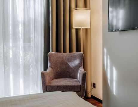 Grauer Sessel neben einer Stehlampe an einem Fenster mit durchsichtigen Vorhängen in einem gemütlichen, sonnendurchfluteten Raum.