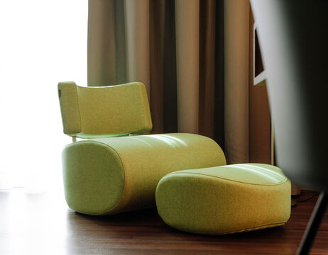 Hellgrüner moderner Sessel mit passender Ottomane auf einem Holzboden, in der Nähe von Vorhängen und weichem Tageslicht.