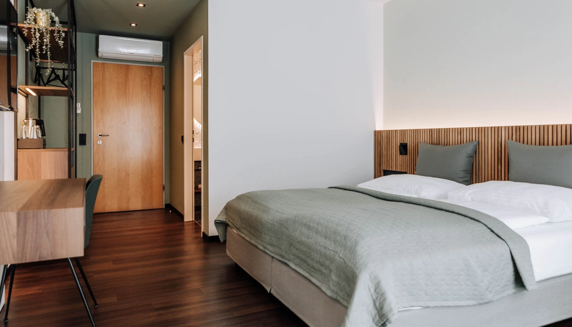 Modernes Hotelzimmer mit sauber gemachtem Bett, Holzboden, Schreibtisch und Tür zum Badezimmer.