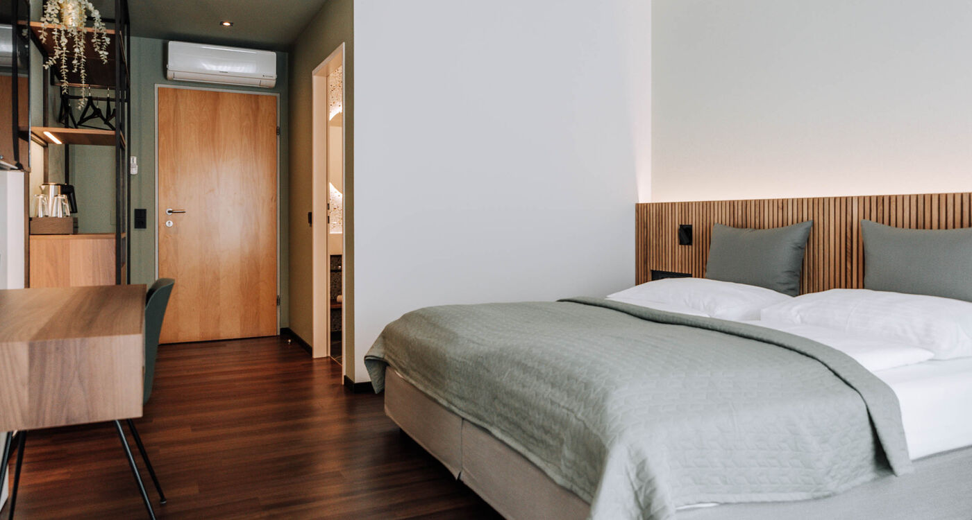 Modern hotel room with neatly made bed, wooden floor, desk and door to the bathroom.
