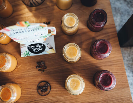 Kleine Gläser mit bunten Säften stehen auf einem Holztisch neben einer teilweise geöffneten bunten Papierschachtel.