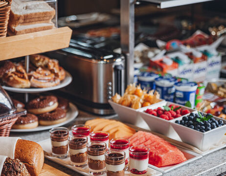 Frühstücksbuffet mit Gebäck, Obst, Miniparfaits, Joghurtbechern und einem Toaster auf einer Arbeitsplatte.