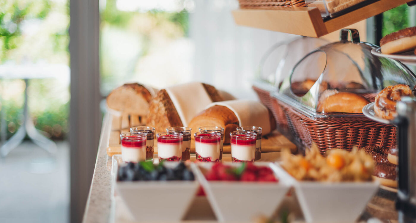 Frühstücksbuffet mit verschiedenen Brotsorten, Gebäck, Joghurtparfaits und Schalen mit frischen Beeren auf einer Theke.
