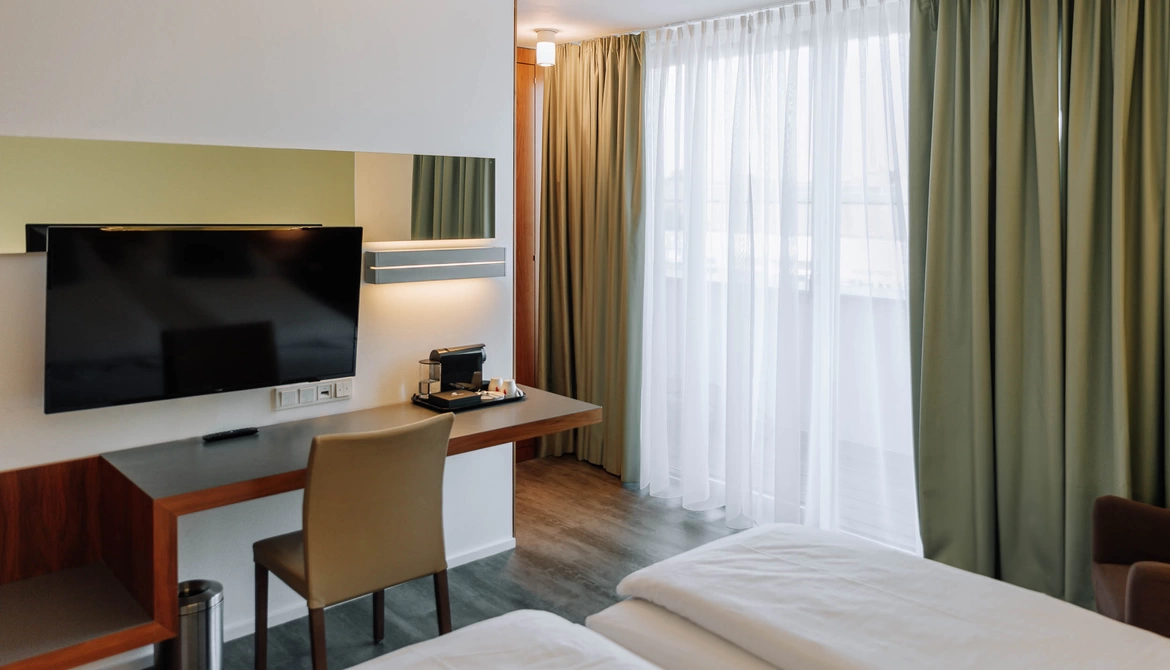 Modernes Hotelzimmer mit Bett, Schreibtisch, TV, Stuhl, Kaffeemaschine und großem Fenster mit grünen Vorhängen.