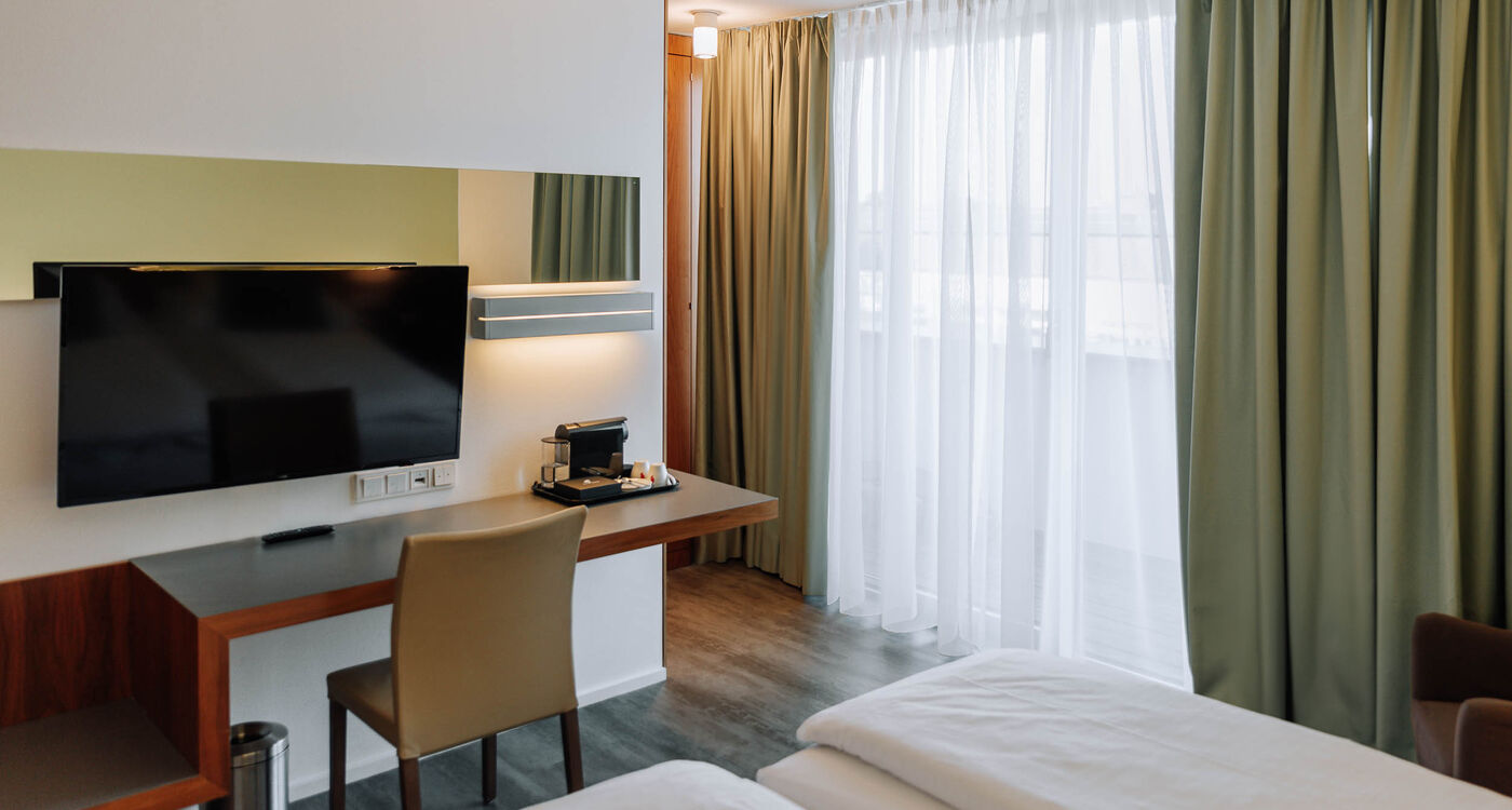 Modern hotel room with bed, desk, TV, chair, coffee machine and large window with green curtains.