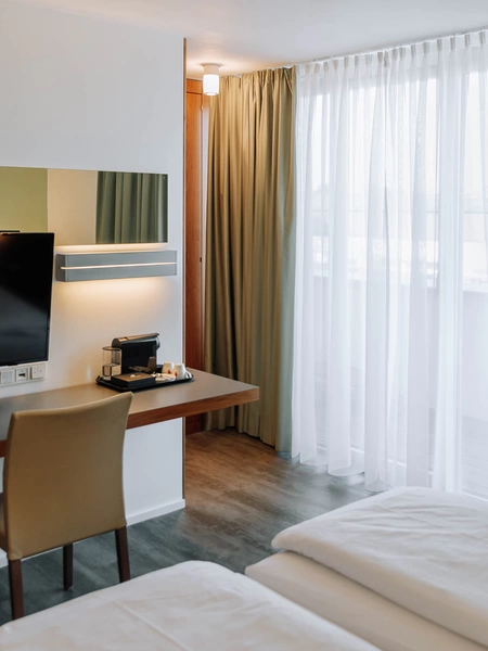 Modern hotel room with bed, desk, TV, chair, coffee machine and large window with green curtains.