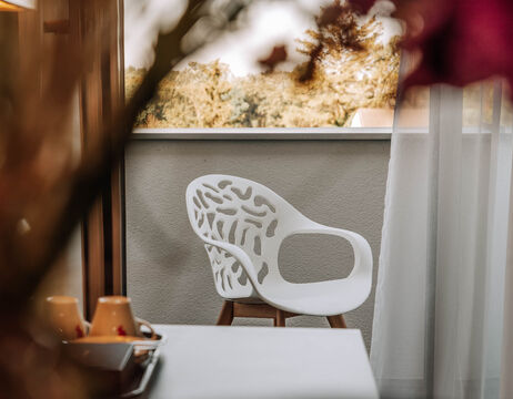 A modern white chair on a balcony near a window, with blurred flowers and cups in the foreground.