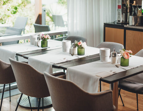 Modernes Café mit ordentlich gedeckten Tischen, weißen Tischläufern, Blumen und weichen grauen Stühlen an großen Fenstern.