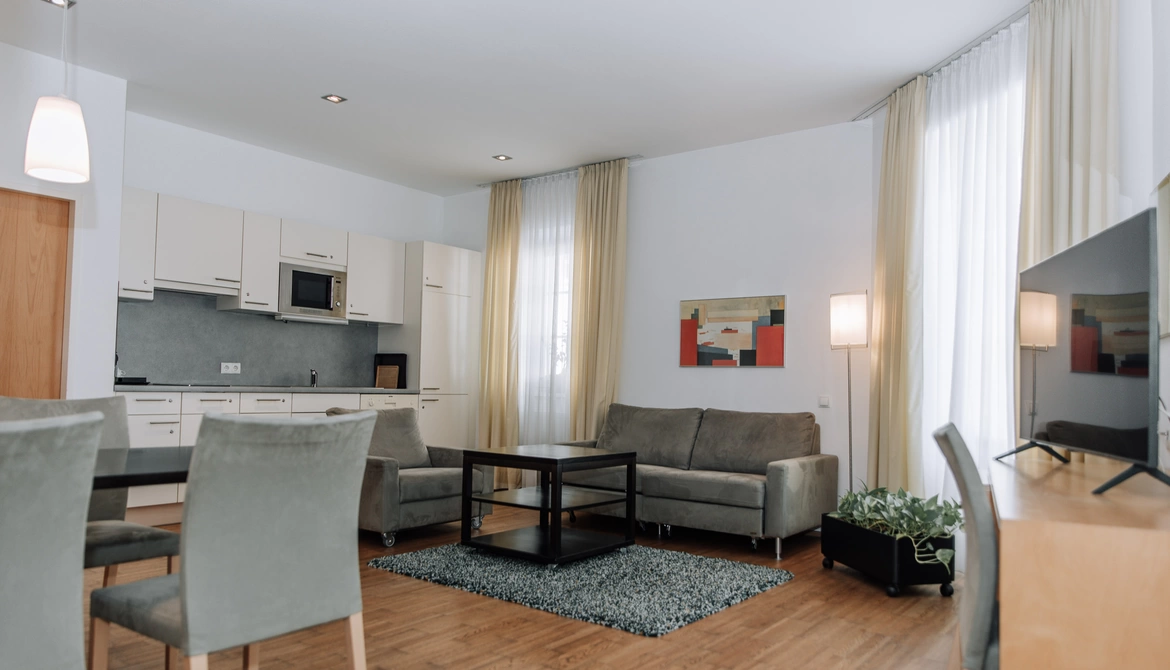 Modernes Wohnzimmer mit grauen Sofas, Esstisch, Küchenzeile, großen Fenstern und hellen Vorhängen.