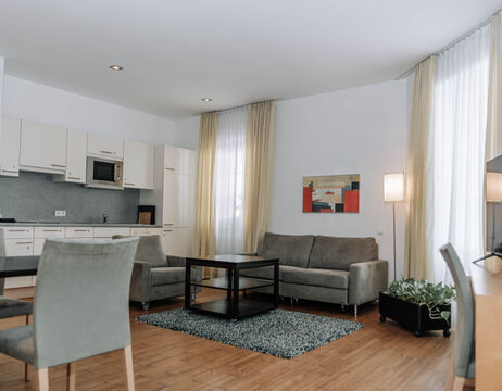Modern living room and kitchen with gray sofas, dining area and large windows with light curtains.