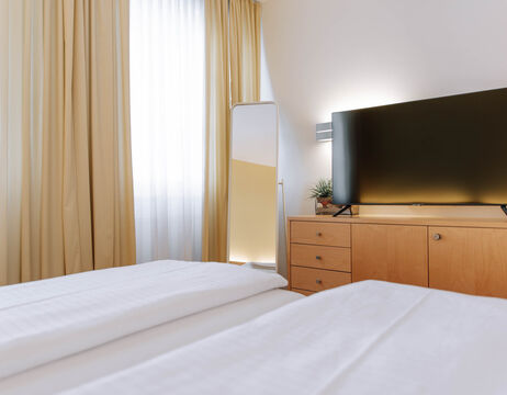 Modern hotel room with a neatly made bed, flat-screen TV on a wooden chest of drawers, mirror and light-colored curtains.