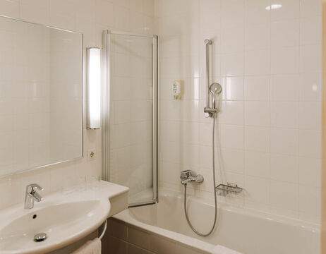A modern bathroom with washbasin, mirror, white tiles and a bathtub with shower head and glass partition.