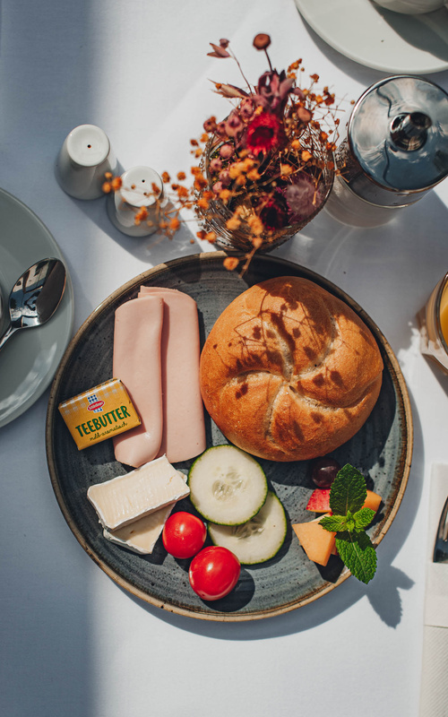 Brötchen, Wurst, Käse, Butter, Gurke, Kirschtomaten und Garnitur auf einem Tisch anrichten.