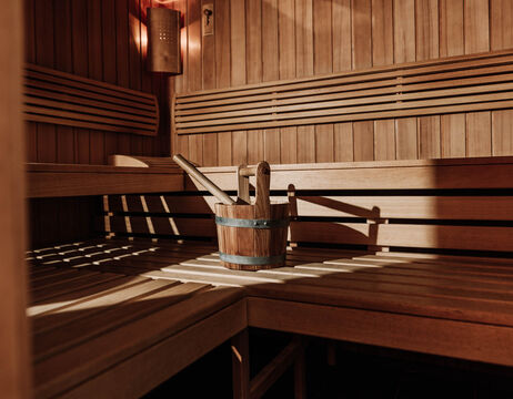 Interior of the wooden sauna with benches, a bucket and a ladle in the middle, bathed in warm light.