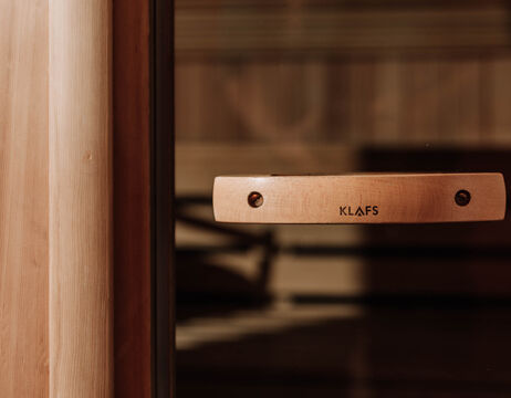 Wooden sauna door handle with engraved word KLAFS, shown in front of a blurred sauna interior.