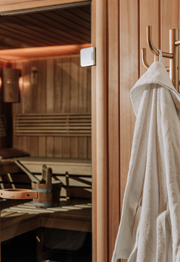A white bathrobe hangs on a hook in front of a wooden sauna with benches and a bucket inside.