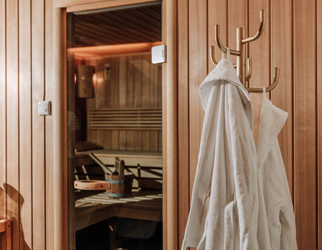 Two white bathrobes hang on a clothes rack in front of a wooden sauna cabin with benches and warm lighting.