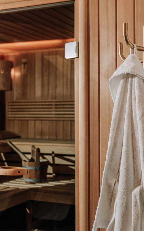 White bathrobe on a hook in front of a wooden sauna cabin with benches and a bucket of water inside.
