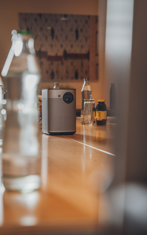 Ein tragbarer Projektor steht auf einem Holztisch, umgeben von Flaschen in einem schwach beleuchteten Raum.