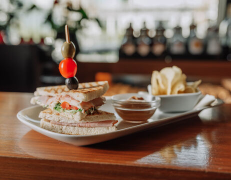 Club sandwich with olives on a skewer, served with chips and dip on a white plate in a restaurant.