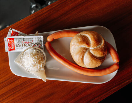 Two sausages, a roll, sauerkraut and mustard packets on a white plate on a wooden table.
