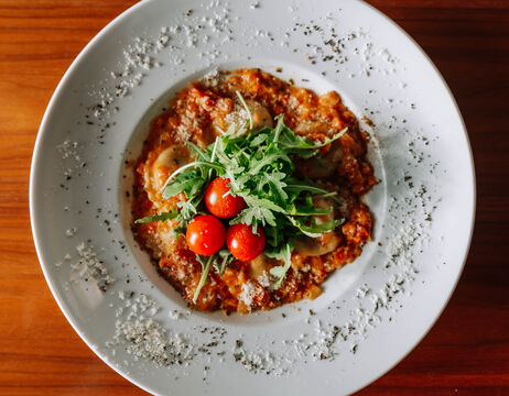 A white plate of ravioli topped with rocket, cherry tomatoes and grated cheese sits on a wooden table.