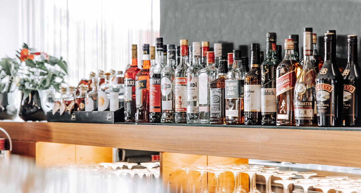 Various bottles of schnapps are lined up on a bar counter, with glassware underneath.