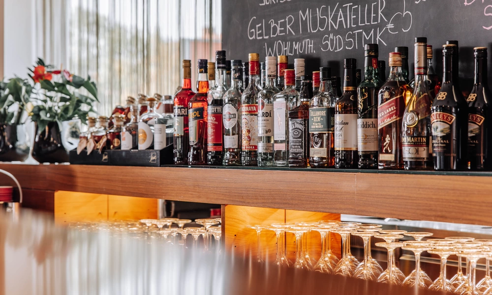 Eine Reihe verschiedener Schnapsflaschen auf einem Barregal mit Gläsern darunter und einer Speisekarte auf einer Kreidetafel darüber.