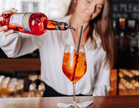 A bartender pours Aperol into a glass with ice and a straw on a bar counter.