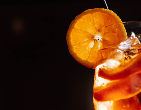 Close-up of an orange slice on a glass filled with ice and an amber-colored drink against a dark background.