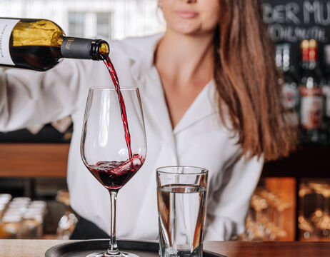 Woman in white shirt pouring red wine into a glass next to a glass of water on a tray at a bar.
