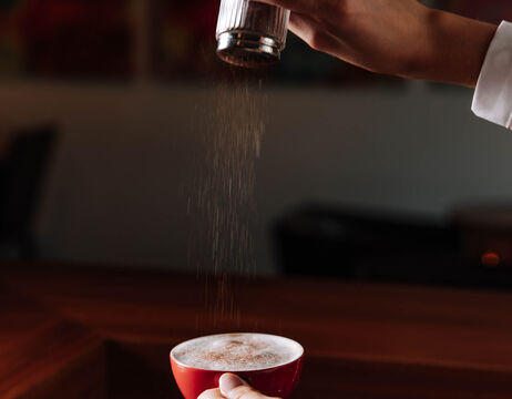 A hand sprinkles cinnamon or cocoa powder on a cup of coffee in a red mug.