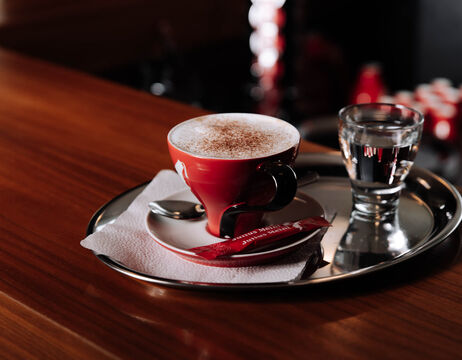 A red cup of cappuccino and a glass of water on a tray, with a chili pepper on the saucer.