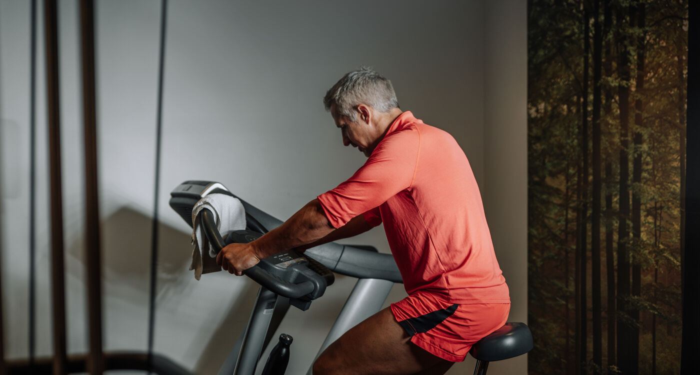 A man in red sportswear is riding a stationary bike in a hall with a towel over the handlebars.
