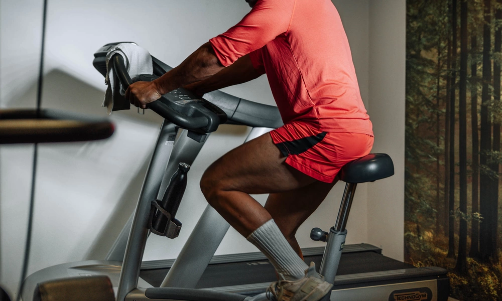 A person in red training clothes is riding a stationary bike in a hall near a wall with a forest mural.
