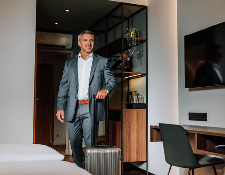 Ein lächelnder Mann im Anzug betritt ein Hotelzimmer mit einem Rollkoffer.