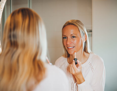 A woman applies lip gloss while looking at herself in the bathroom mirror.