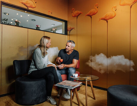 Zwei Menschen sitzen und unterhalten sich in einem Raum mit Flamingo-Wandbildern, einem kleinen Tisch und Zeitschriften.