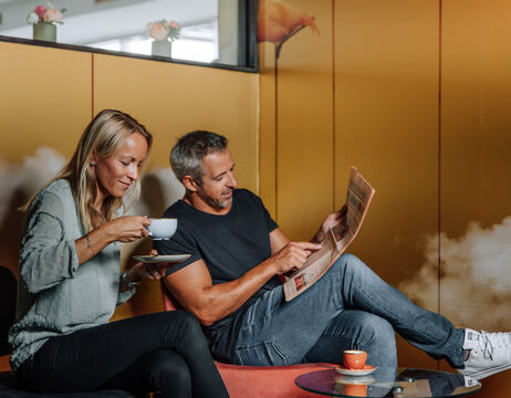 Eine Frau, die Kaffee trinkt, und ein Mann, der eine Zeitung liest, sitzen zusammen in einem gemütlichen, modernen Raum.