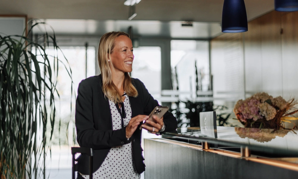 Lächelnde Frau in Geschäftskleidung, die an einer modernen Rezeption ihr Telefon benutzt.
