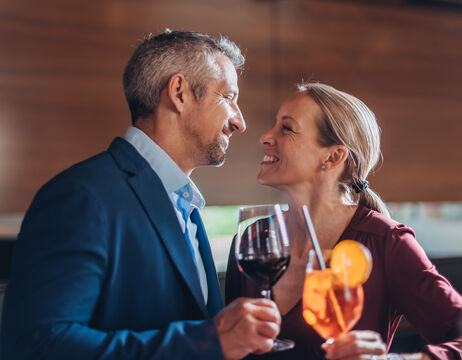 A smiling couple toast with wine and a cocktail while leaning close together in a restaurant.