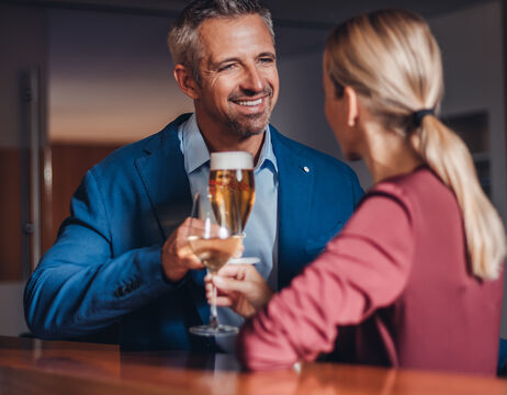 A smiling man and a smiling woman clink glasses of beer and wine at a bar counter.