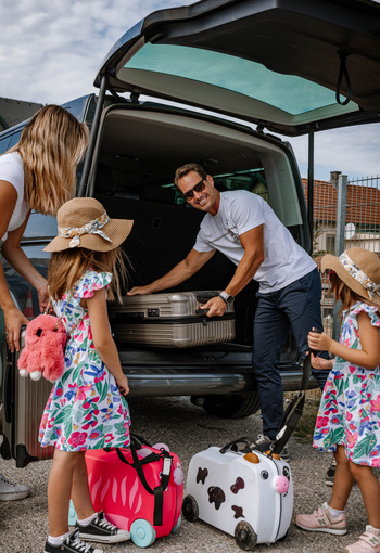 Family loading luggage into a car; two girls with hats and suitcases, adults helping in the back.