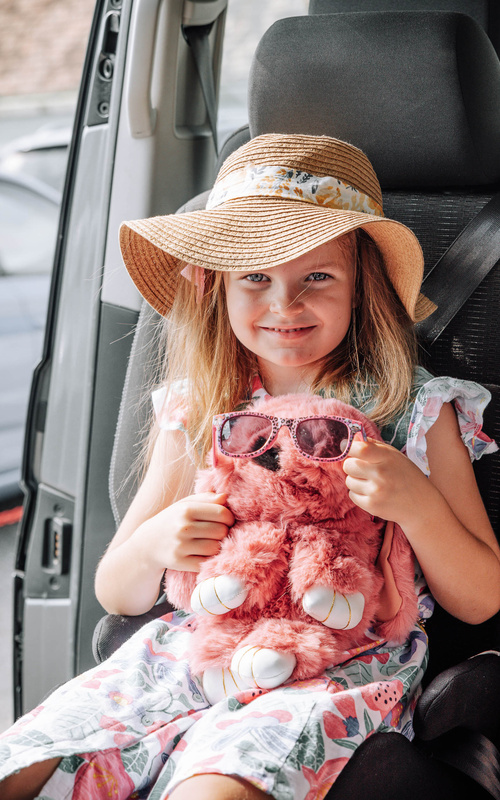 Lächelndes Mädchen mit Sonnenhut hält ein rosafarbenes Stofftier mit Sonnenbrille, während es in einem Autositz sitzt.